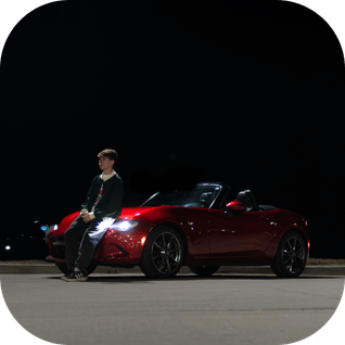 Young man leaning against a red convertible sports car parked on a dark night street.