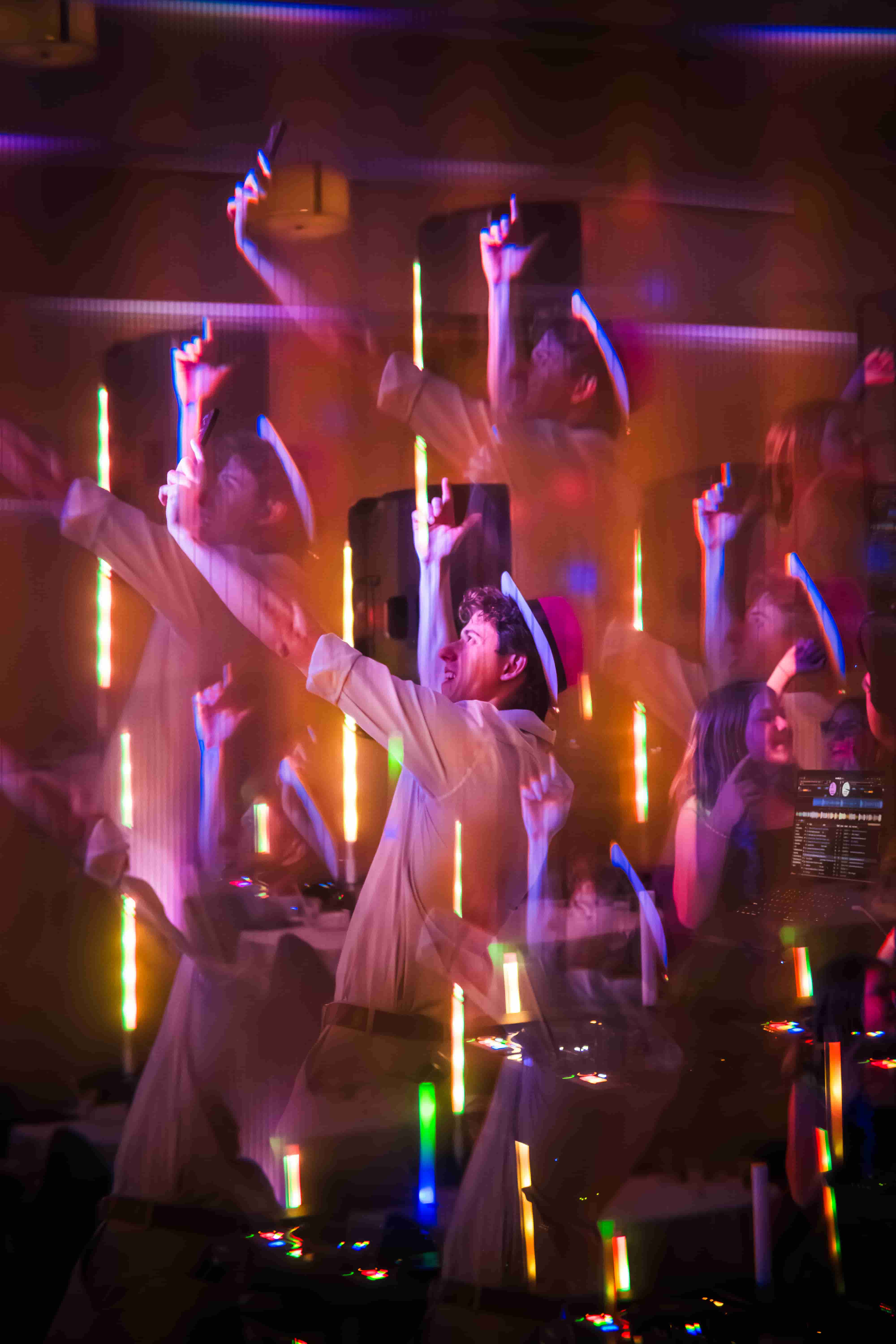 A man in a hat dancing with arms raised under colorful lights at a party with a DJ setup.