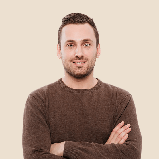 Smiling man with short brown hair wearing a brown sweater with arms crossed against a beige background.