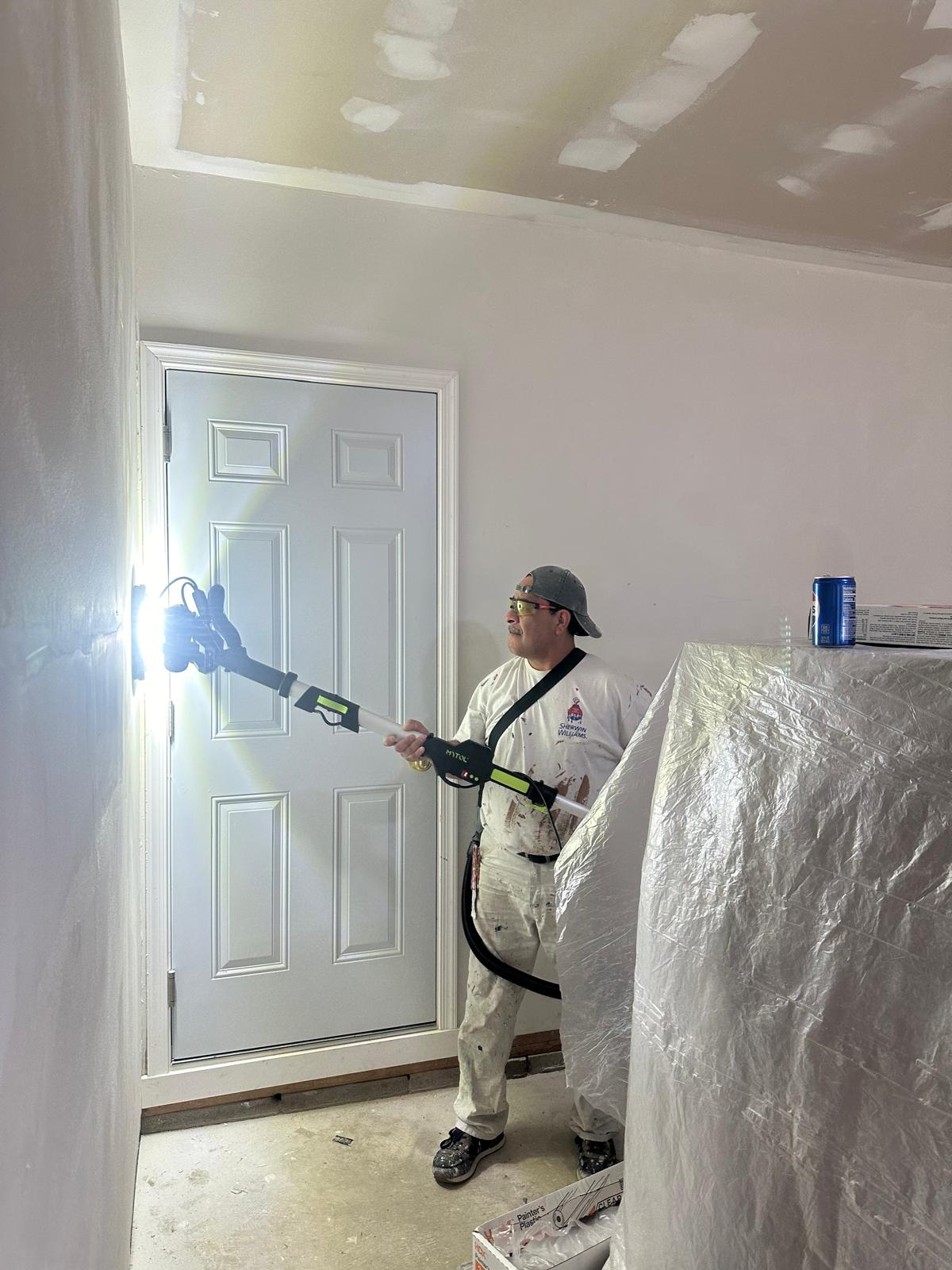 Painter in white clothes using a lighted paint roller extension to paint a wall near a white door in an unfinished room.