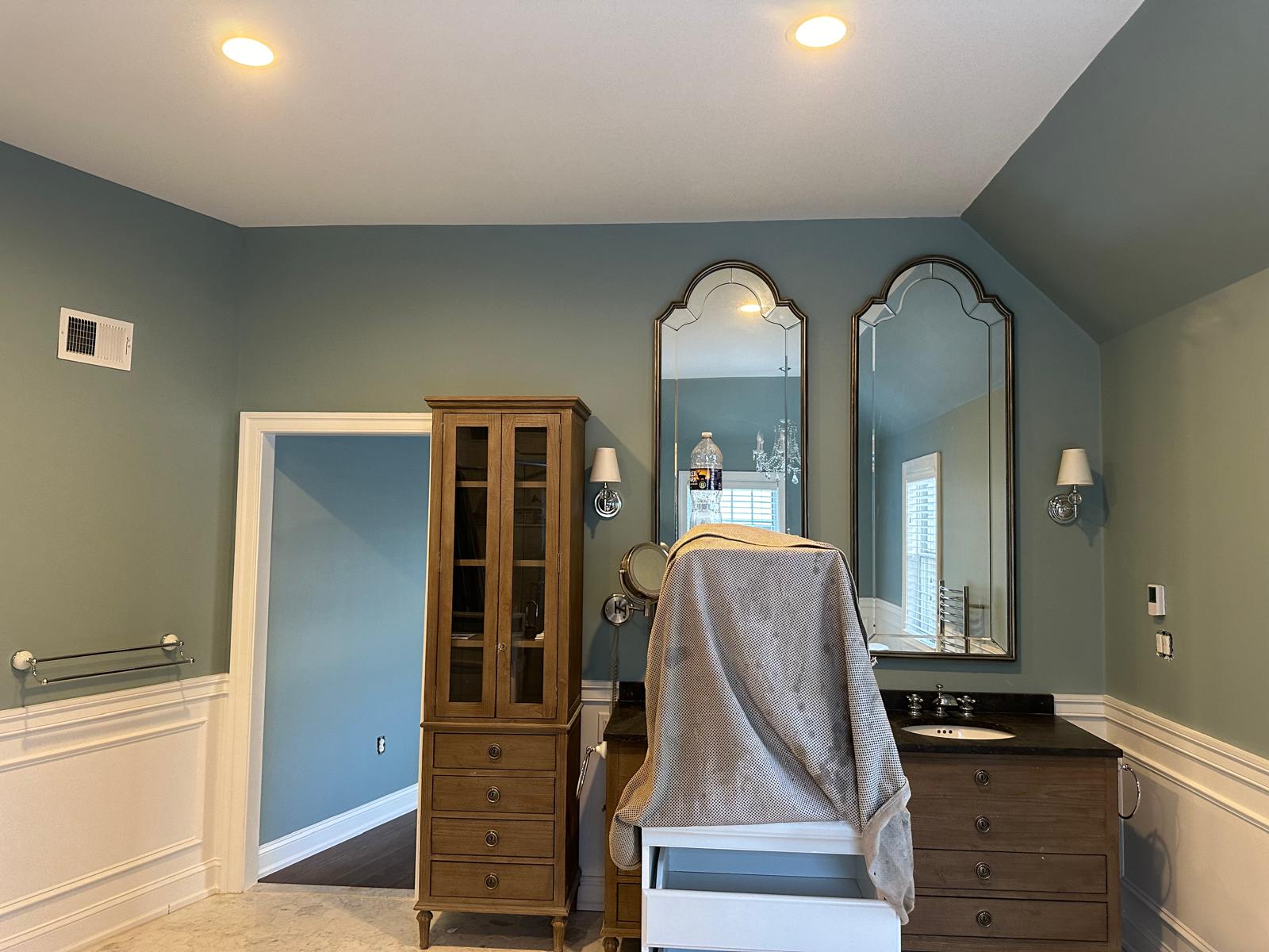 Bathroom with teal walls, tall wooden cabinet, two arched mirrors above a double-sink vanity, and a chair covered with a cloth in the center.