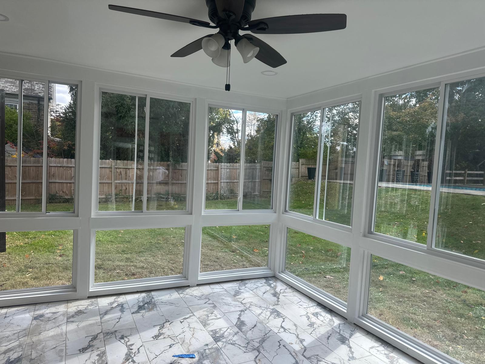 Empty sunroom with large windows on three sides, marble-patterned tile floor, and black ceiling fan with lights.