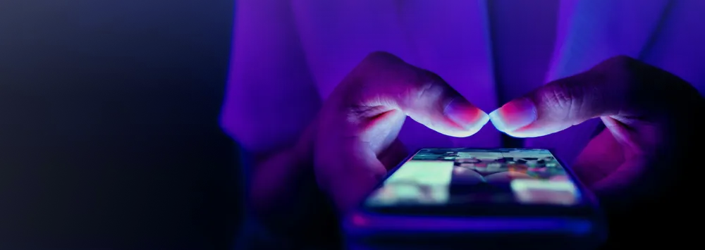 Close-up of hands using a smartphone illuminated by blue and purple light in a dark environment.