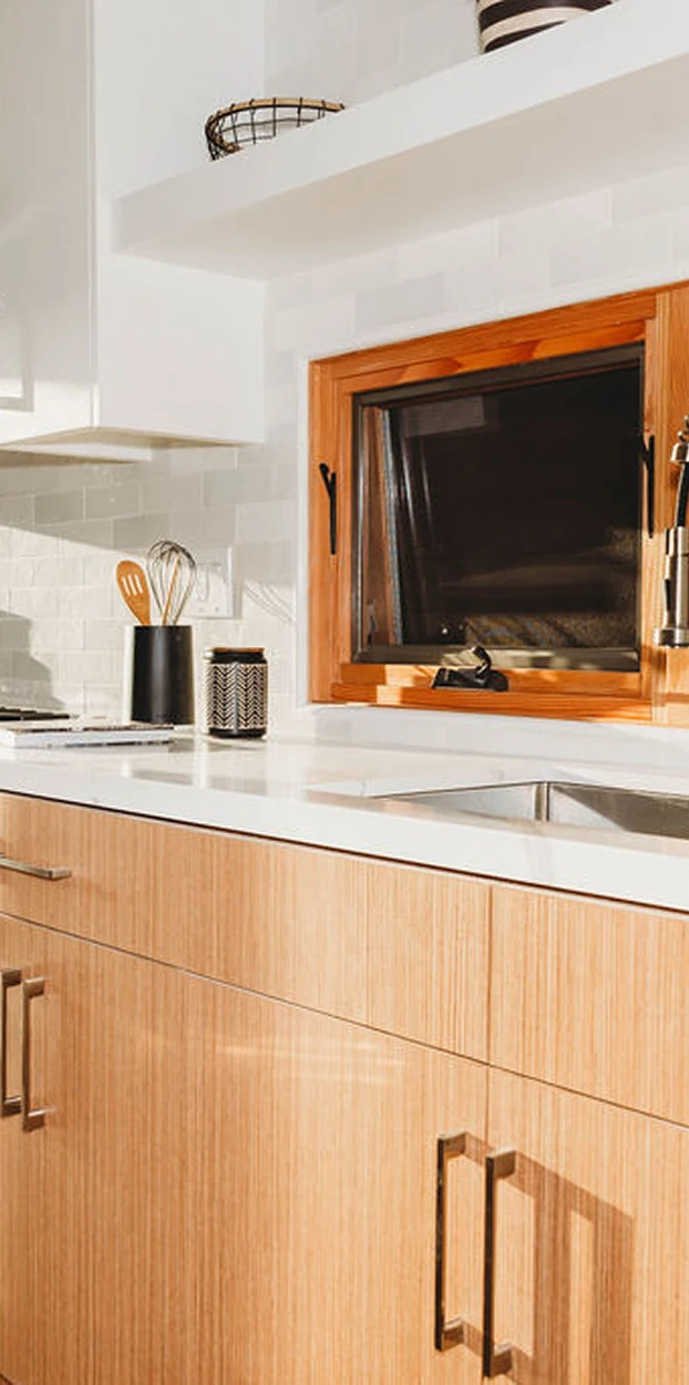 Modern kitchen countertop with light wood cabinets, white backsplash, small utensils holder, and a wooden-framed window above the sink.