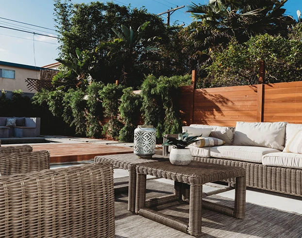 Outdoor patio with wicker furniture including a sofa, armchairs, and coffee table, surrounded by greenery and a wooden fence.