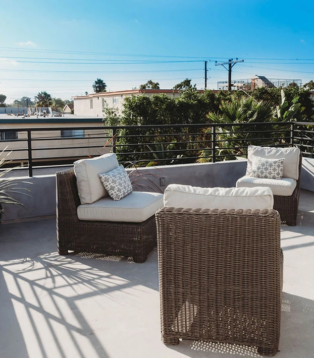 Outdoor patio with three wicker chairs featuring white cushions and patterned pillows under clear blue sky.