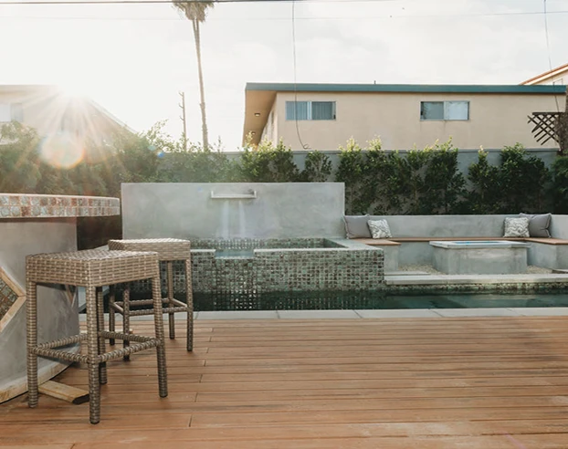 Modern outdoor patio with wooden deck, woven bar stools, built-in seating, and tiled water feature under sunlight.