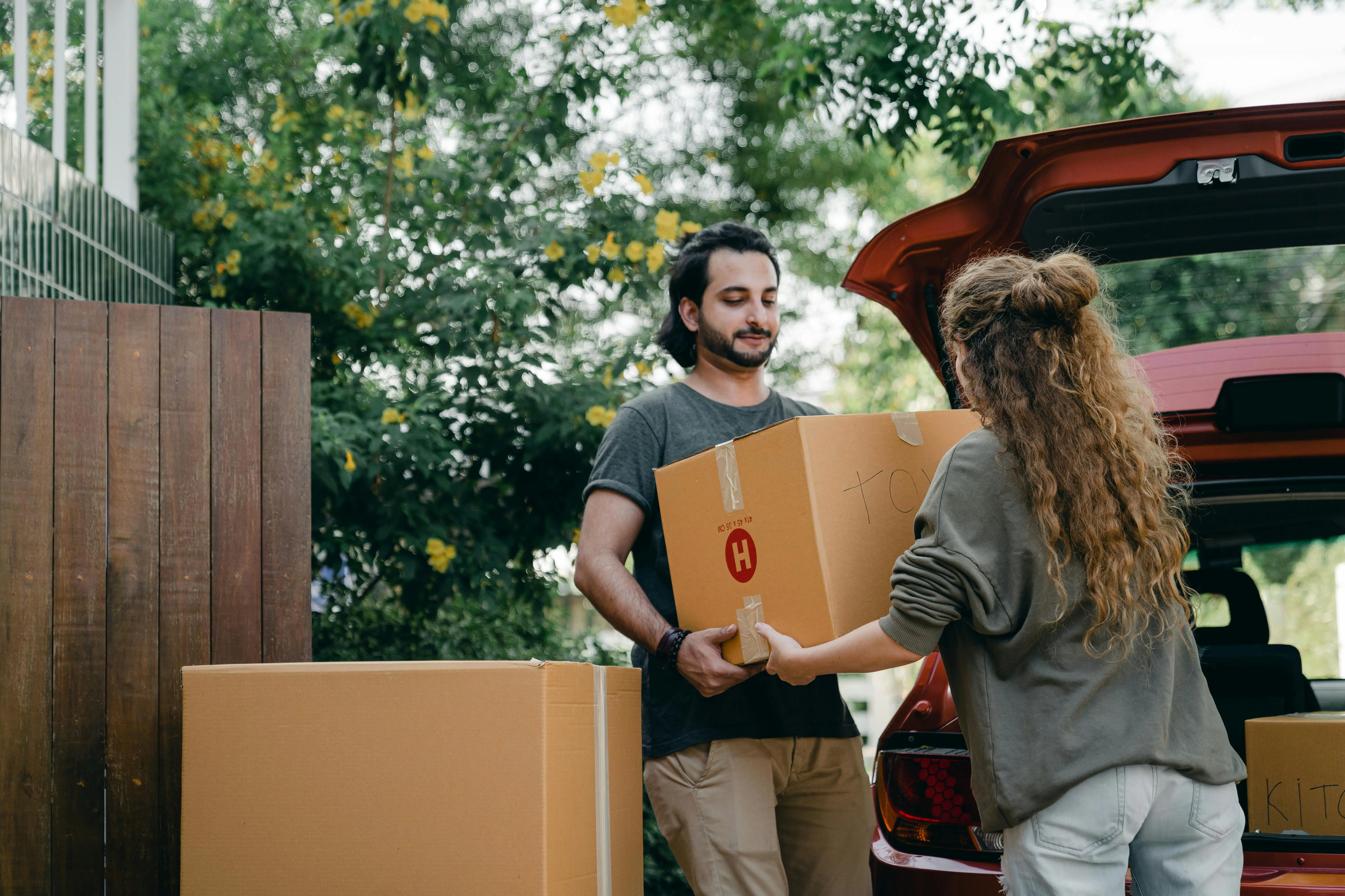 A man and woman carrying cardboard boxes from the back of a red car, surrounded by greenery.