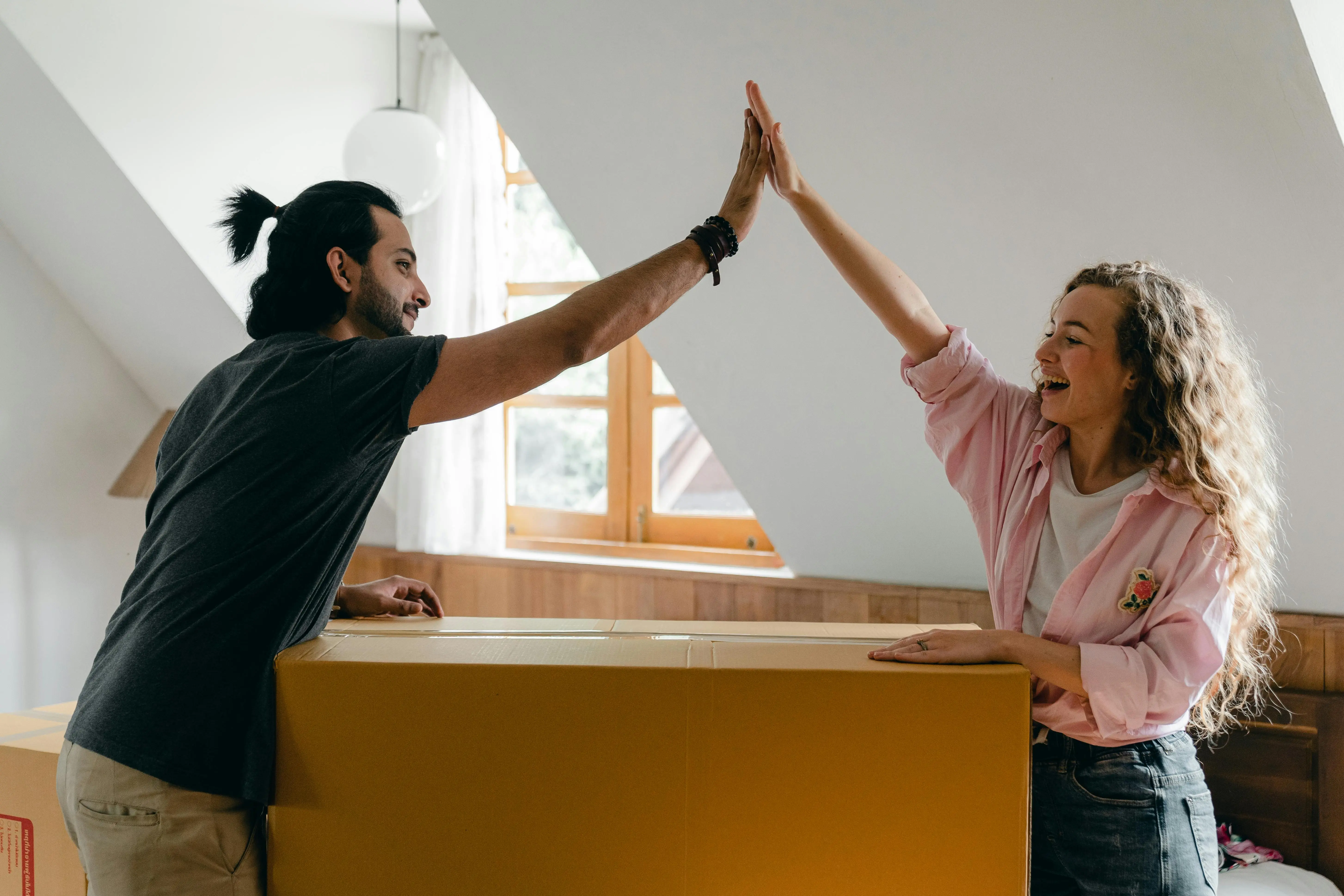 Happy couple high-fiving over a large cardboard box inside a bright room with a window.