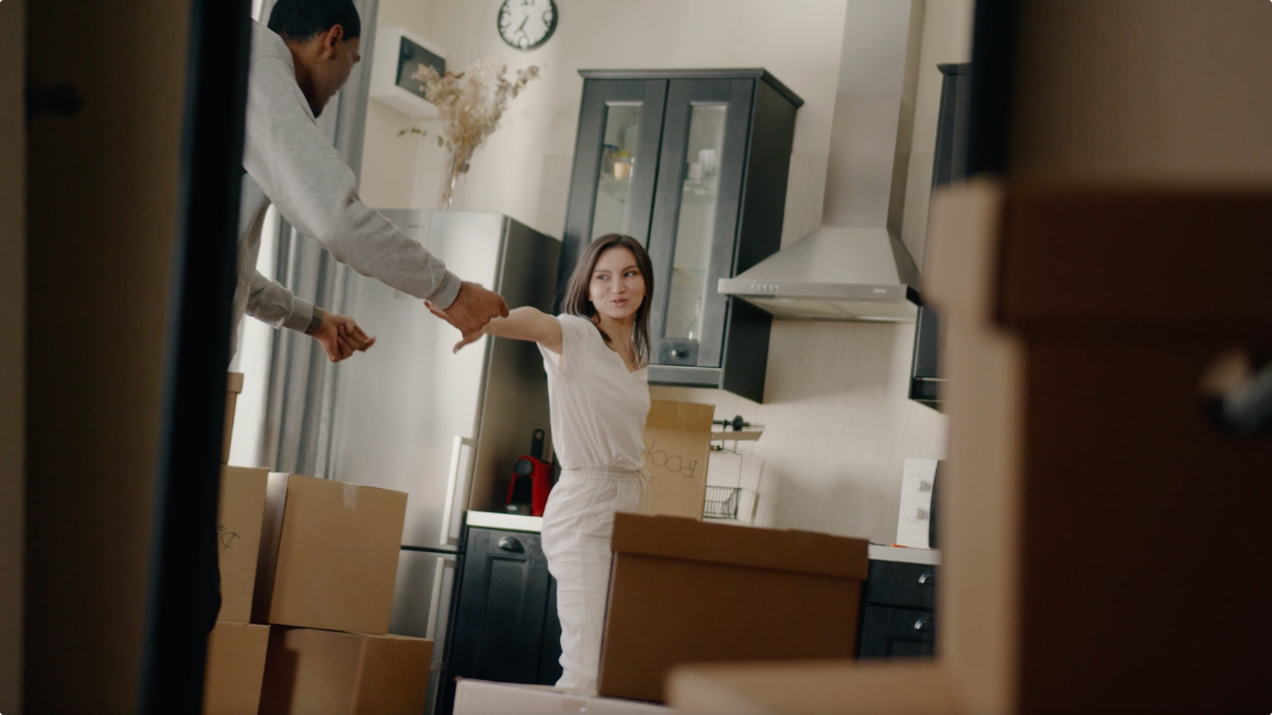 Couple holding hands and smiling in a kitchen surrounded by moving boxes.