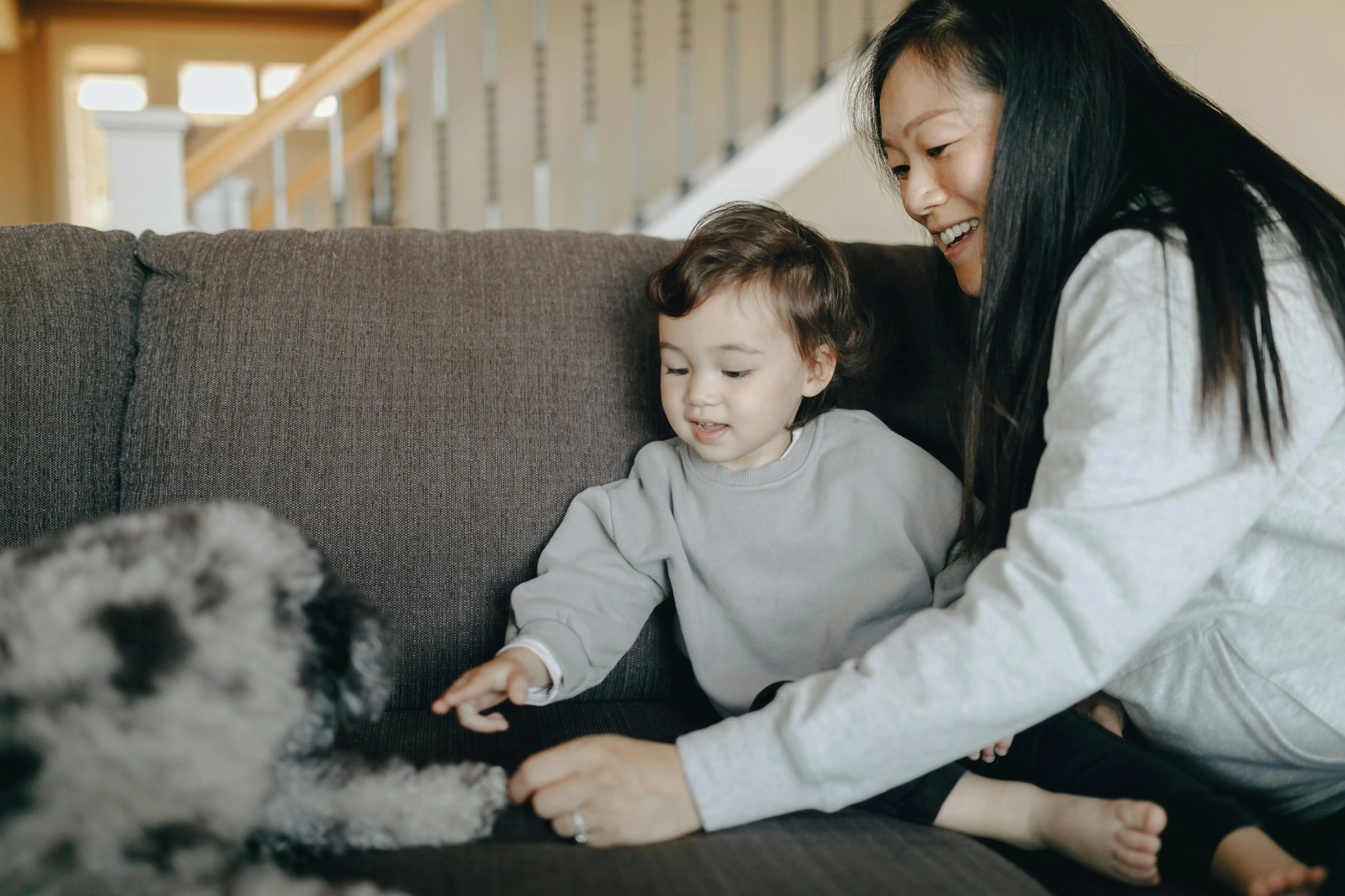 Smiling woman and toddler sitting on a couch reaching out to a fluffy dog.