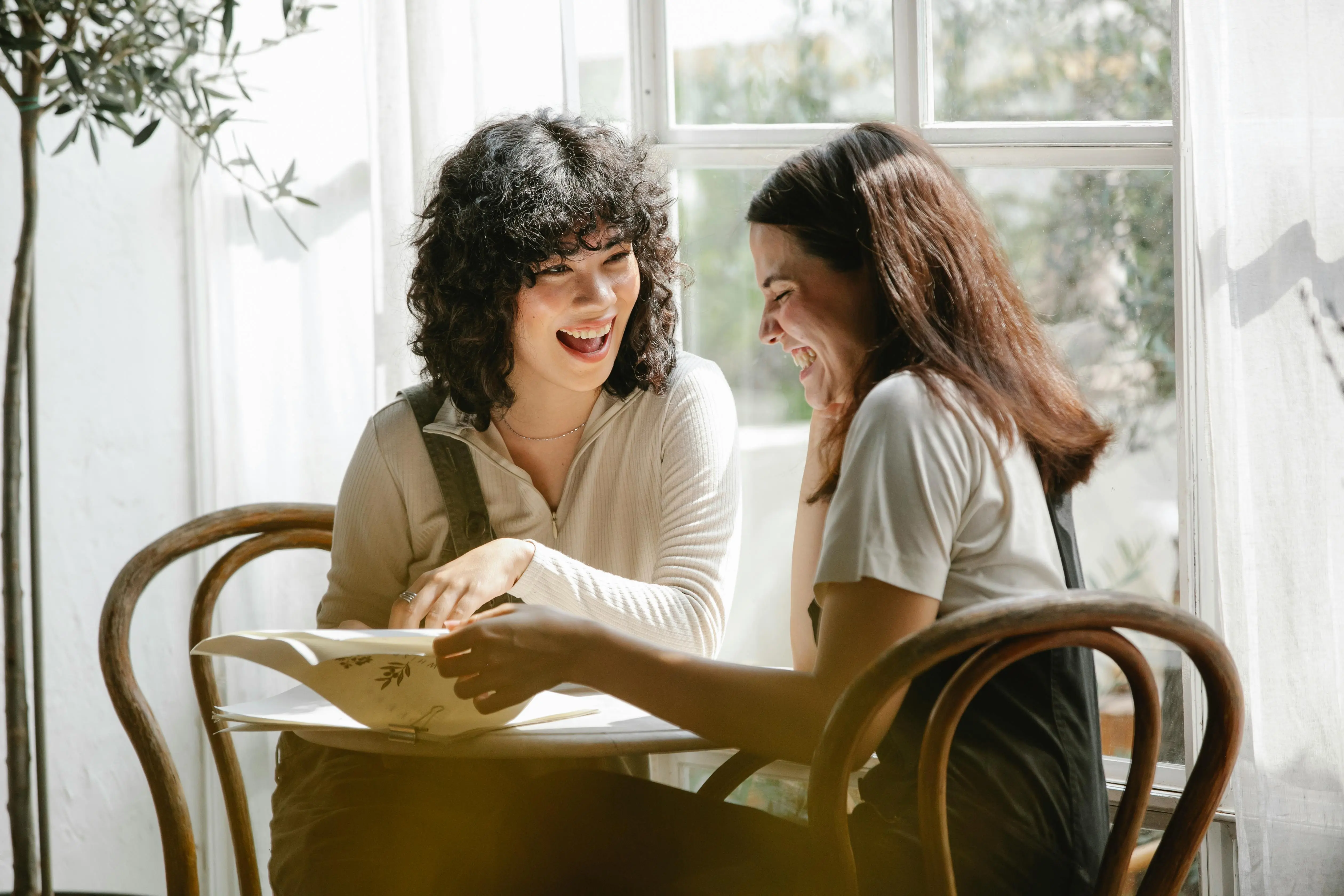 Two young women sitting at a table by a sunny window, laughing and looking at an open book together.
