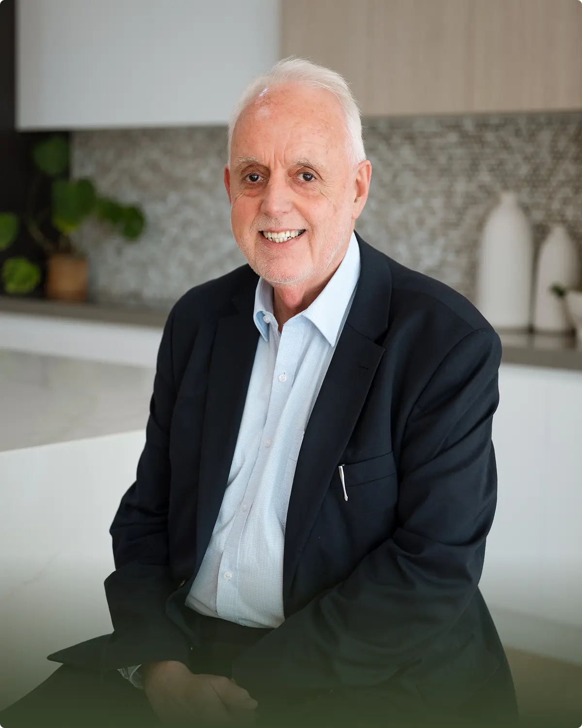 Smiling elderly man with white hair wearing a dark blazer and light blue shirt sitting indoors.