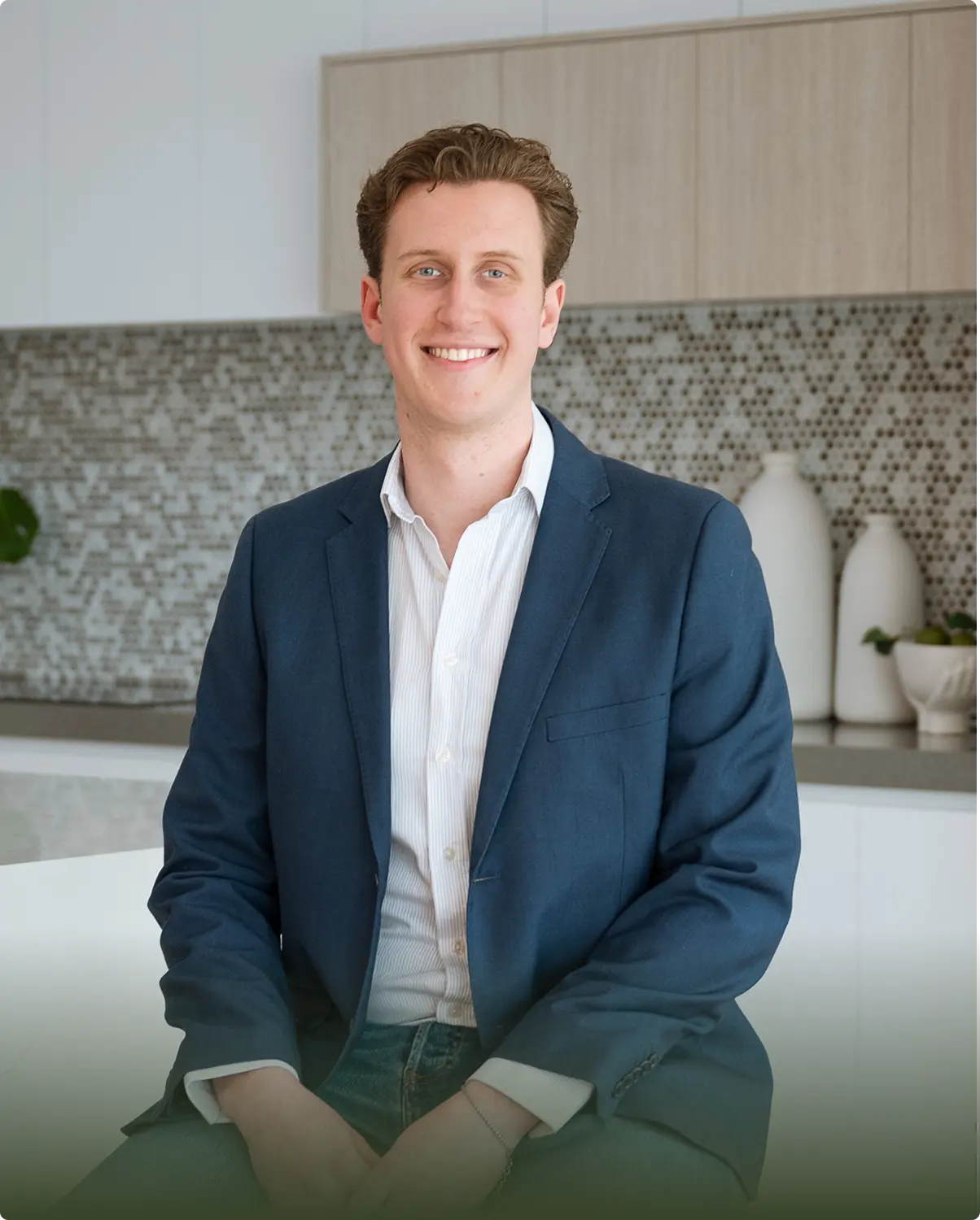 Smiling man with curly brown hair wearing a navy blazer and white shirt sitting in a modern kitchen.