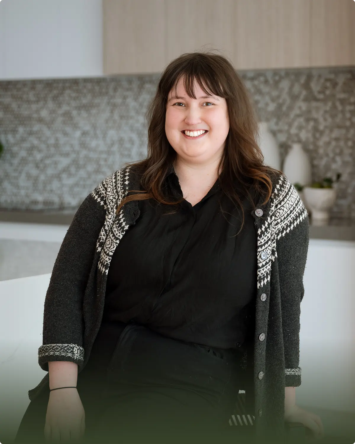 Smiling woman with shoulder-length brown hair wearing a patterned cardigan and black shirt, sitting indoors.