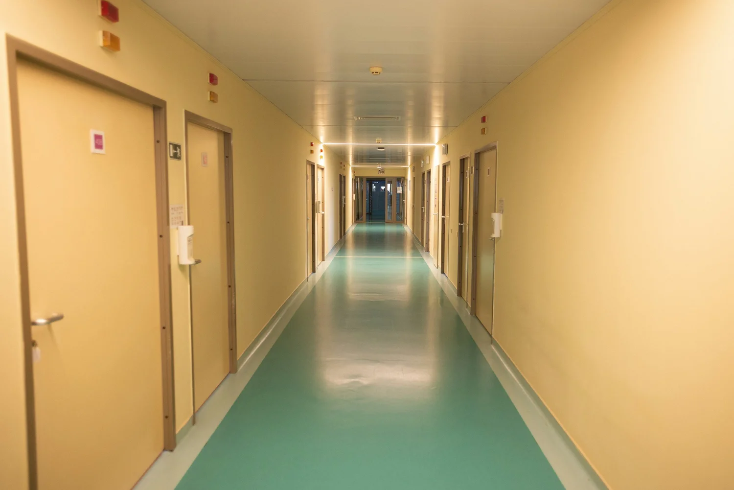 Empty hospital hallway with closed beige doors on both sides and a teal floor.