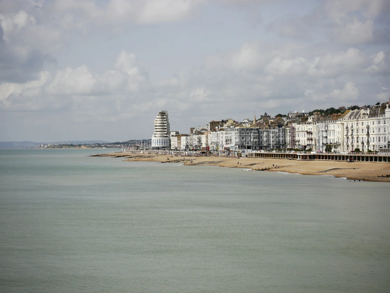 Brighton coast with sandy beaches, a distinctive cylindrical white building, and rows of multi-story seafront houses under a partly cloudy sky.