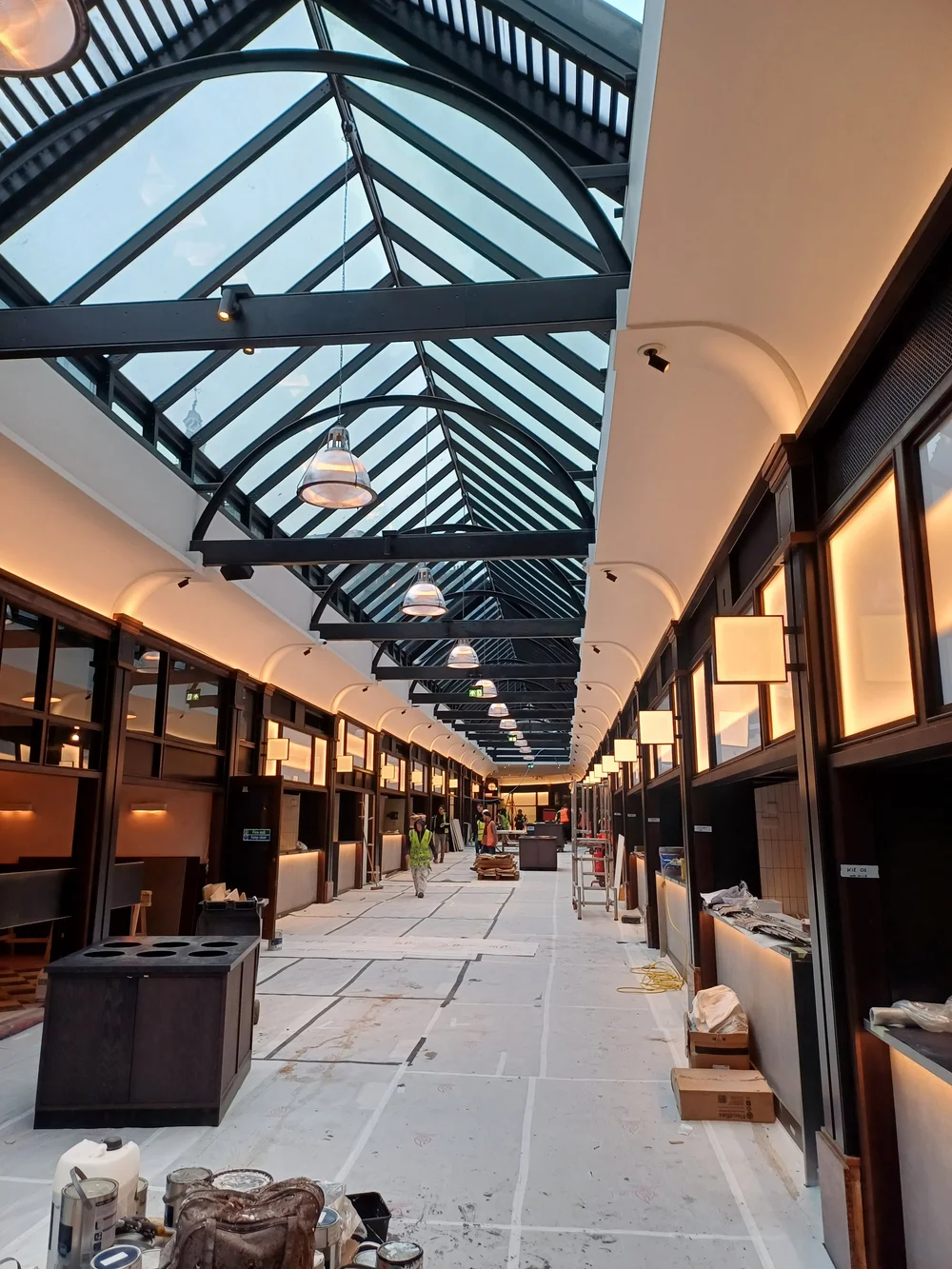 Interior of a large hallway under renovation with a glass ceiling and hanging lights, workers in safety vests, and construction materials on the floor.