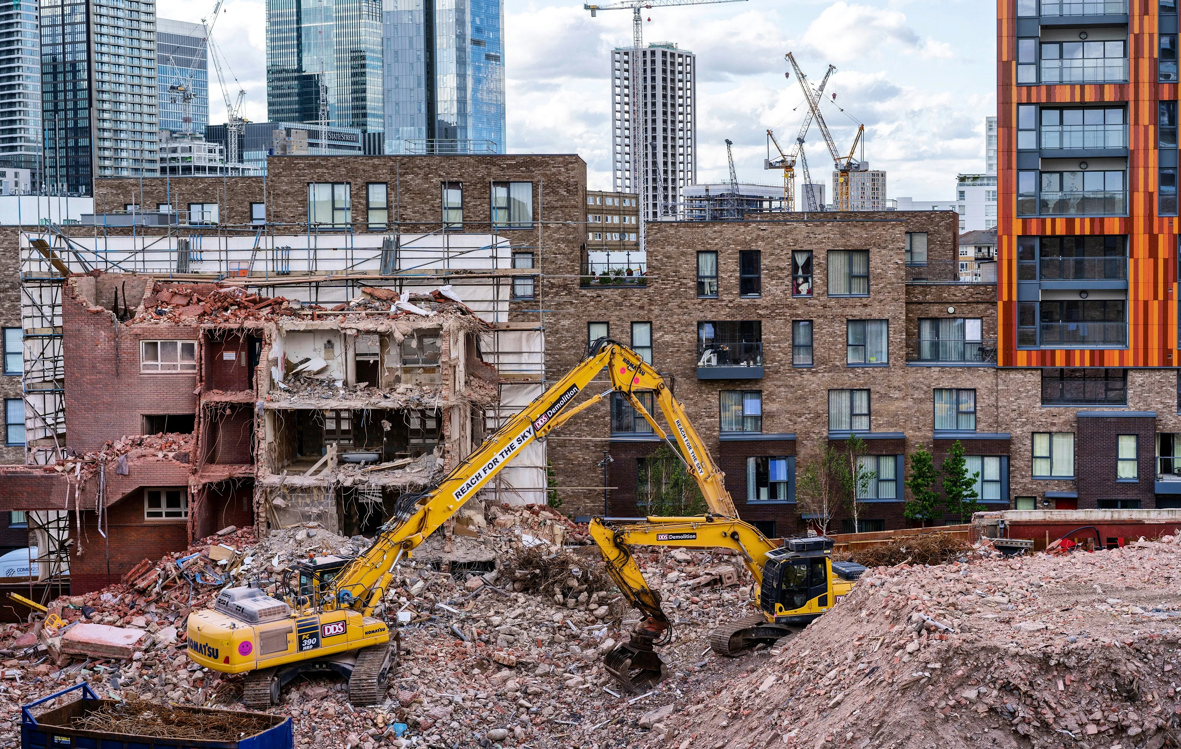 Two yellow excavators demolishing a partially destroyed building amidst rubble with city skyscrapers in the background.