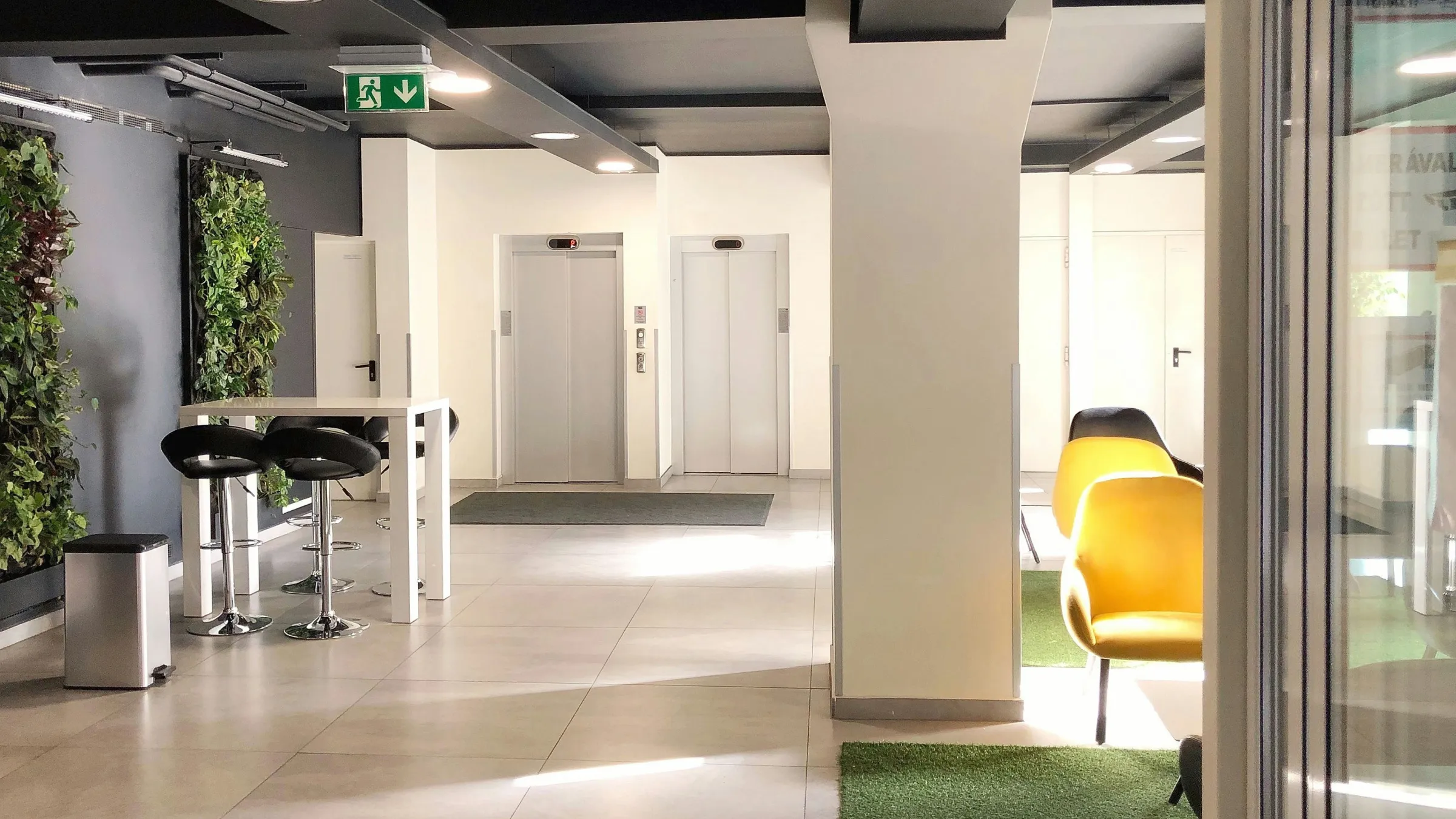 Modern office lobby with two elevators, high white table with black chairs, green vertical plant walls, and yellow lounge chairs on green carpet.