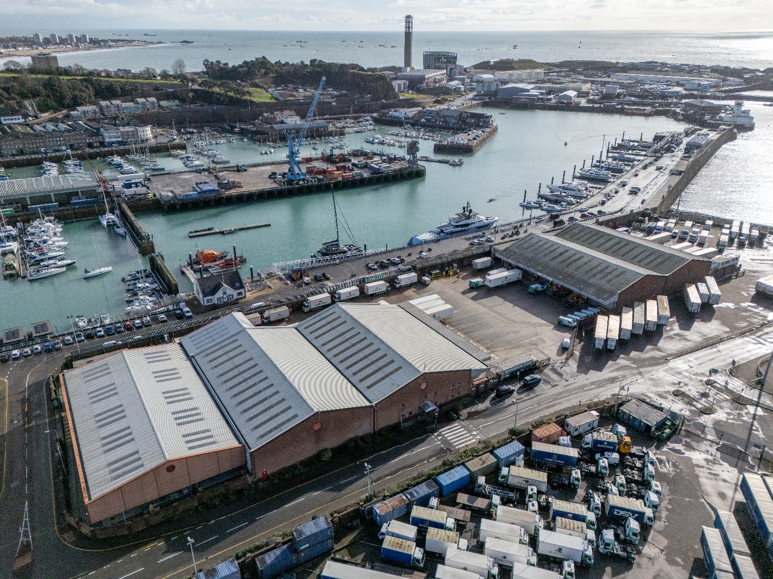 Aerial view of Jersey CI harbor with large warehouses, docked yachts and boats, trucks, and storage containers near the water.