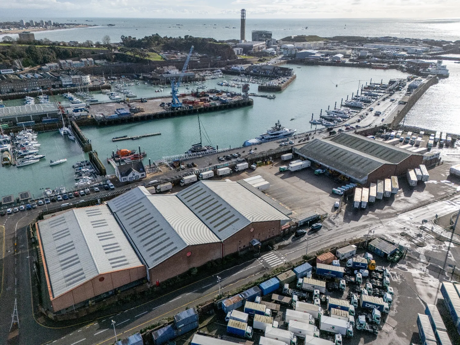 Aerial view of Jersey CI harbor with large warehouses, docked yachts and boats, trucks, and storage containers near the water.