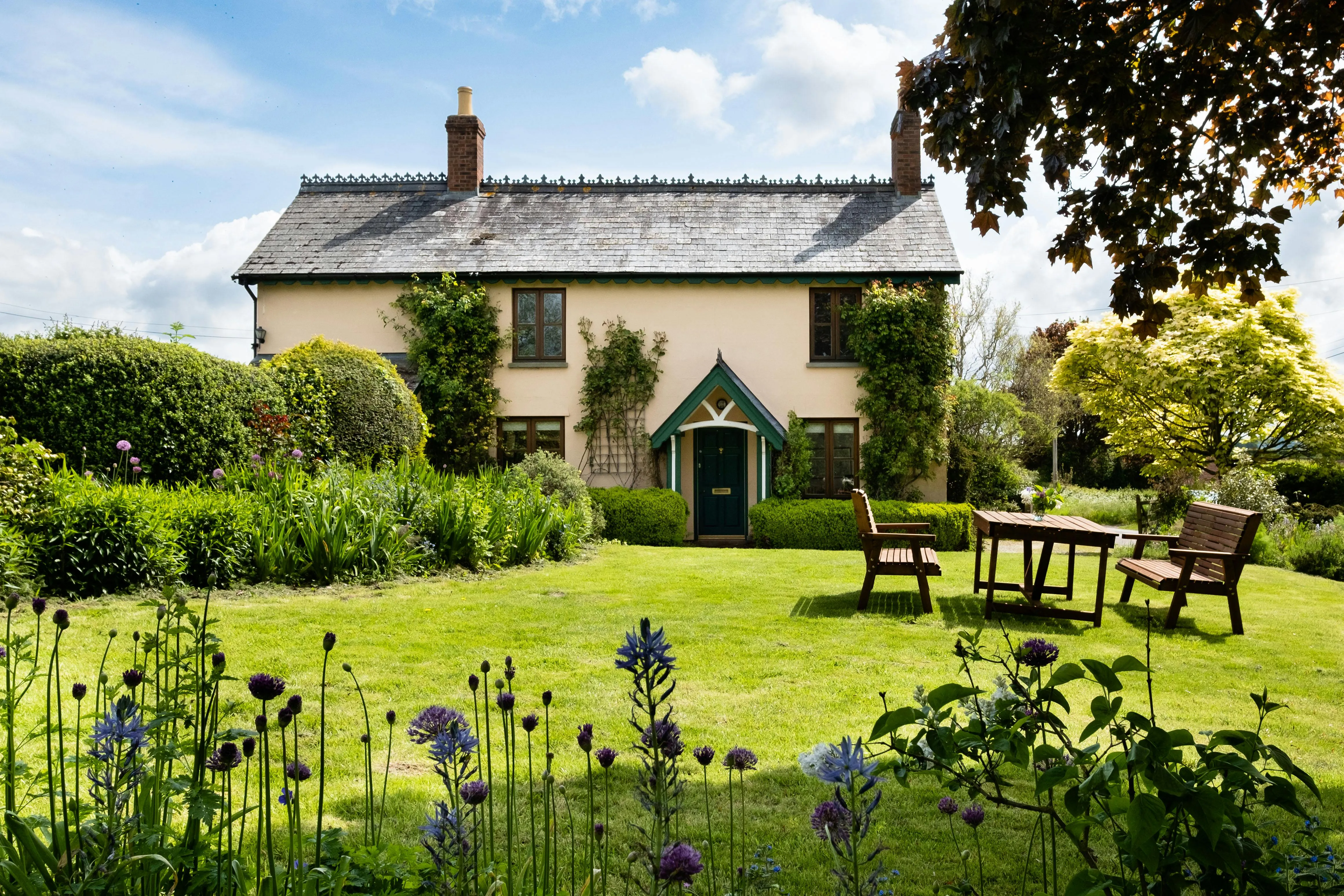 Two-story cottage with a slate roof, green door, surrounded by a lush garden with flowering plants and wooden benches.