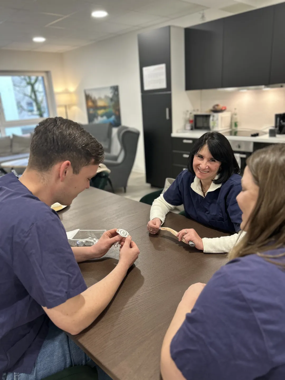 Drei Pflegekräfte reden und lachen am Tisch in einem modernen Aufenthaltsraum