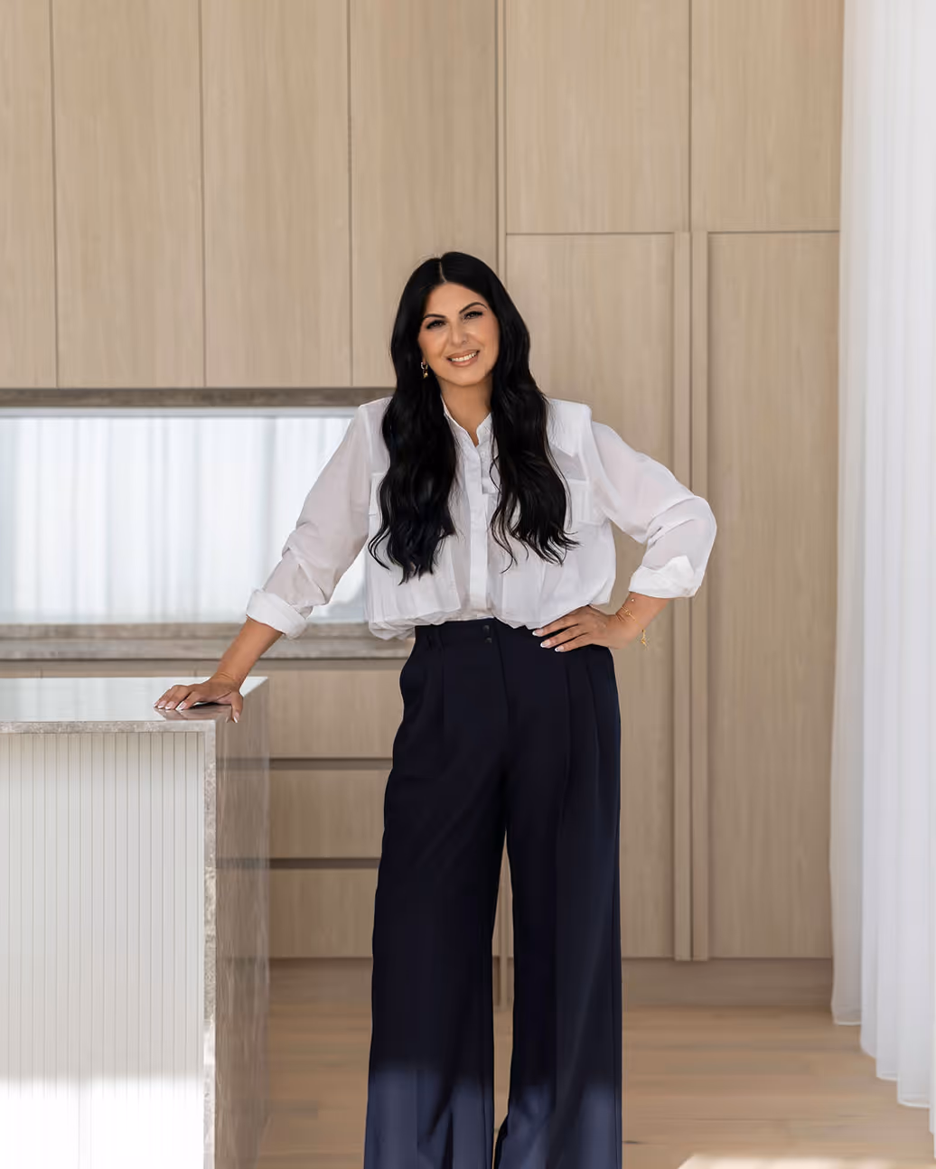 Woman with long black hair wearing a white blouse and black wide-leg pants, leaning on a kitchen island with light wood cabinetry in the background.