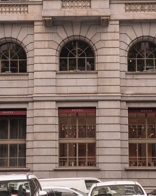 Facade of a stone building with large arched and rectangular windows showcasing a Gucci store, with several vehicles parked in front.