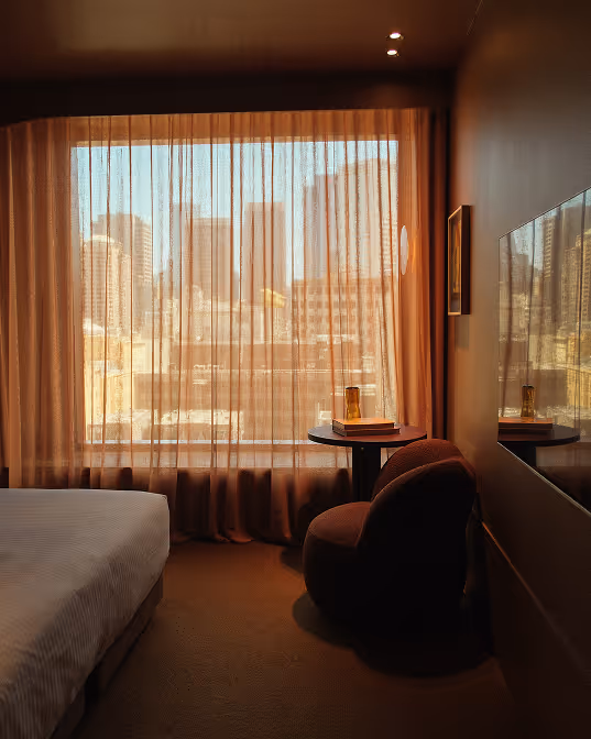 Cozy hotel room with a bed, round chair, small table with books and glass, and a large window with sheer curtains showing a city skyline.
