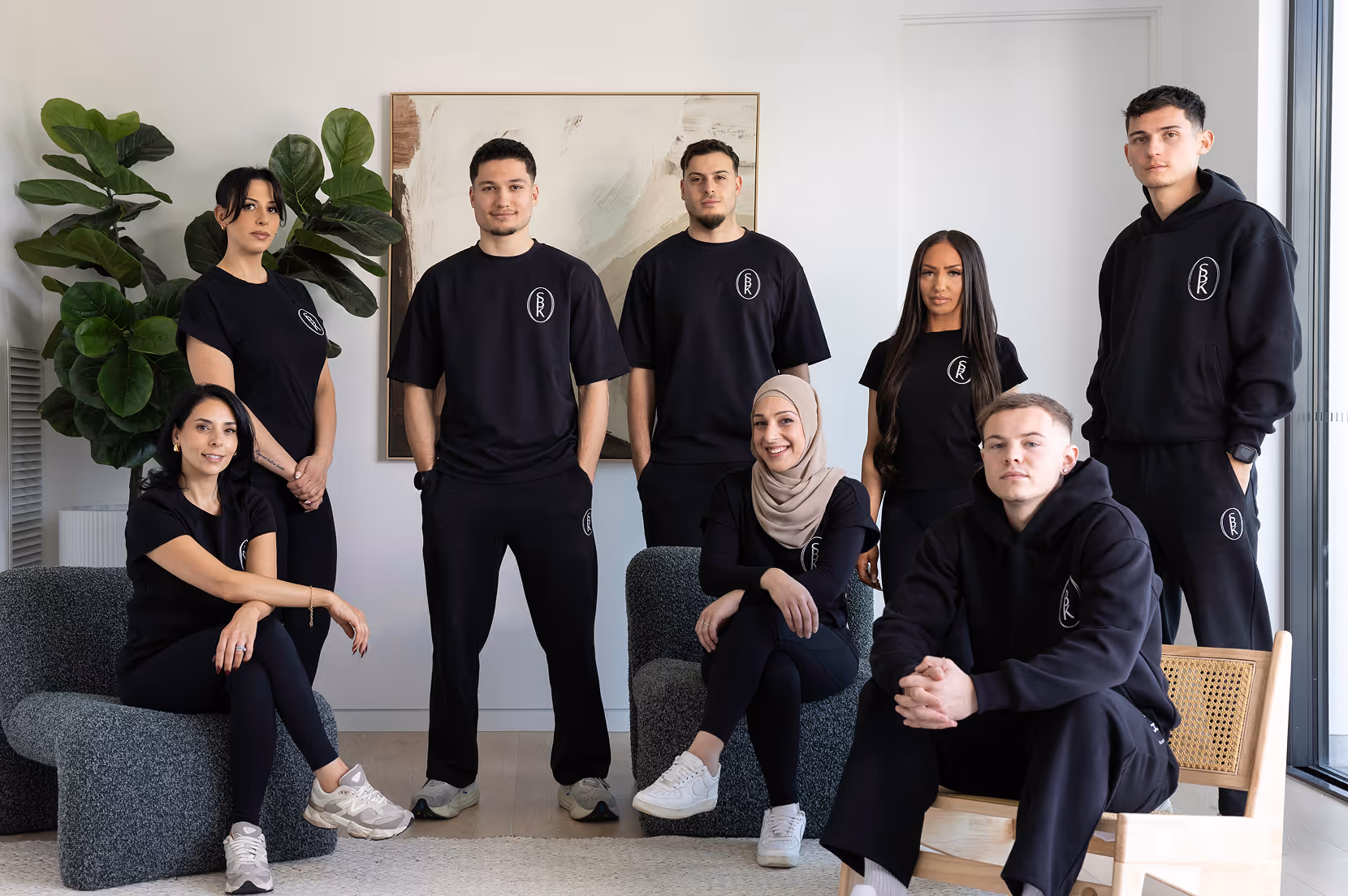 Group of eight diverse young adults wearing matching black casual outfits posing in a modern room with plants and abstract art.