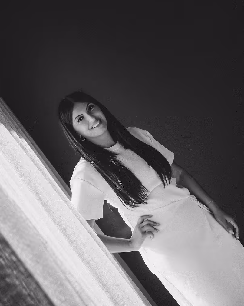 Smiling woman with long dark hair leaning by a window with sheer curtains in a light-colored dress, black and white photo.