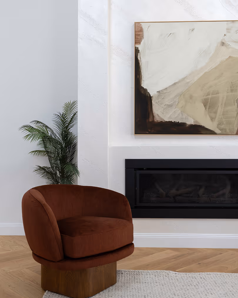 Modern living room corner with a rust-colored upholstered chair, abstract framed artwork above a built-in black fireplace, and a green potted plant.
