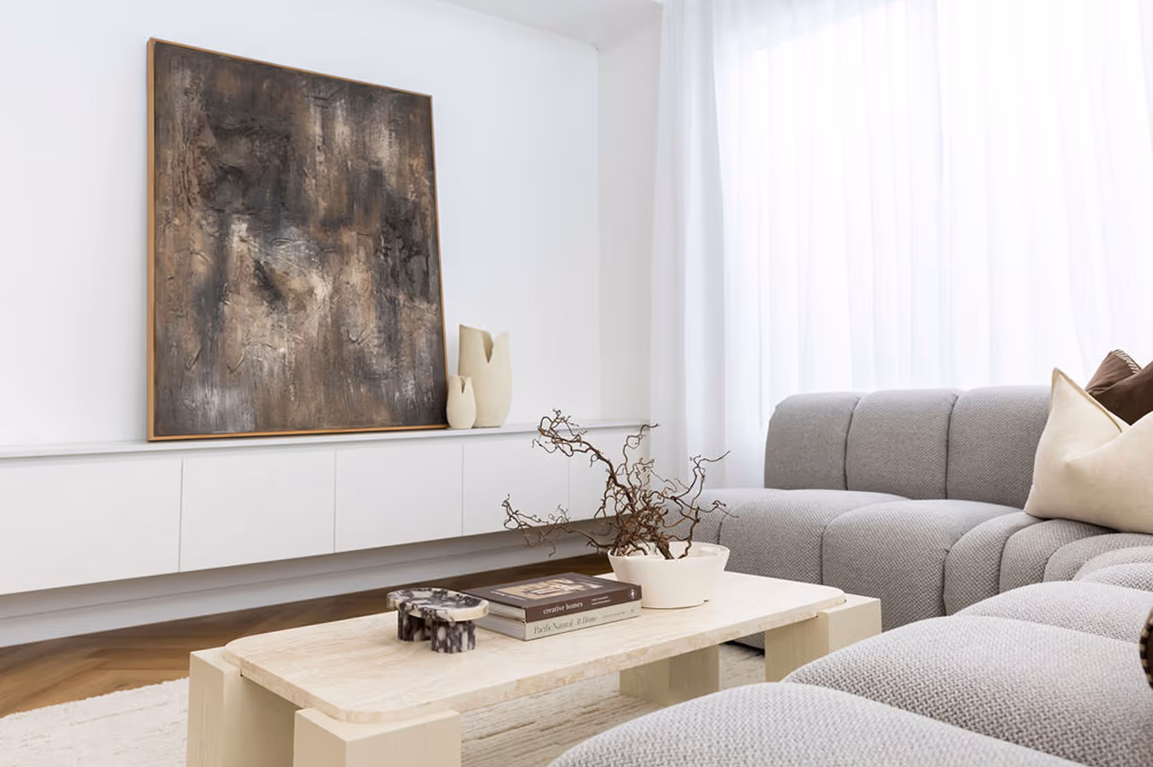 Modern living room with a gray sectional sofa, beige pillows, a stone coffee table with decorative books and a vase with dried branches, and a large abstract painting on a white cabinet against a white wall.