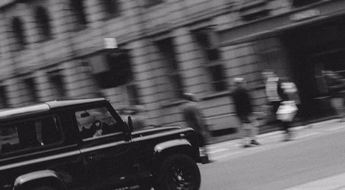 Blurred black and white street scene with a parked black SUV and pedestrians walking on the sidewalk.