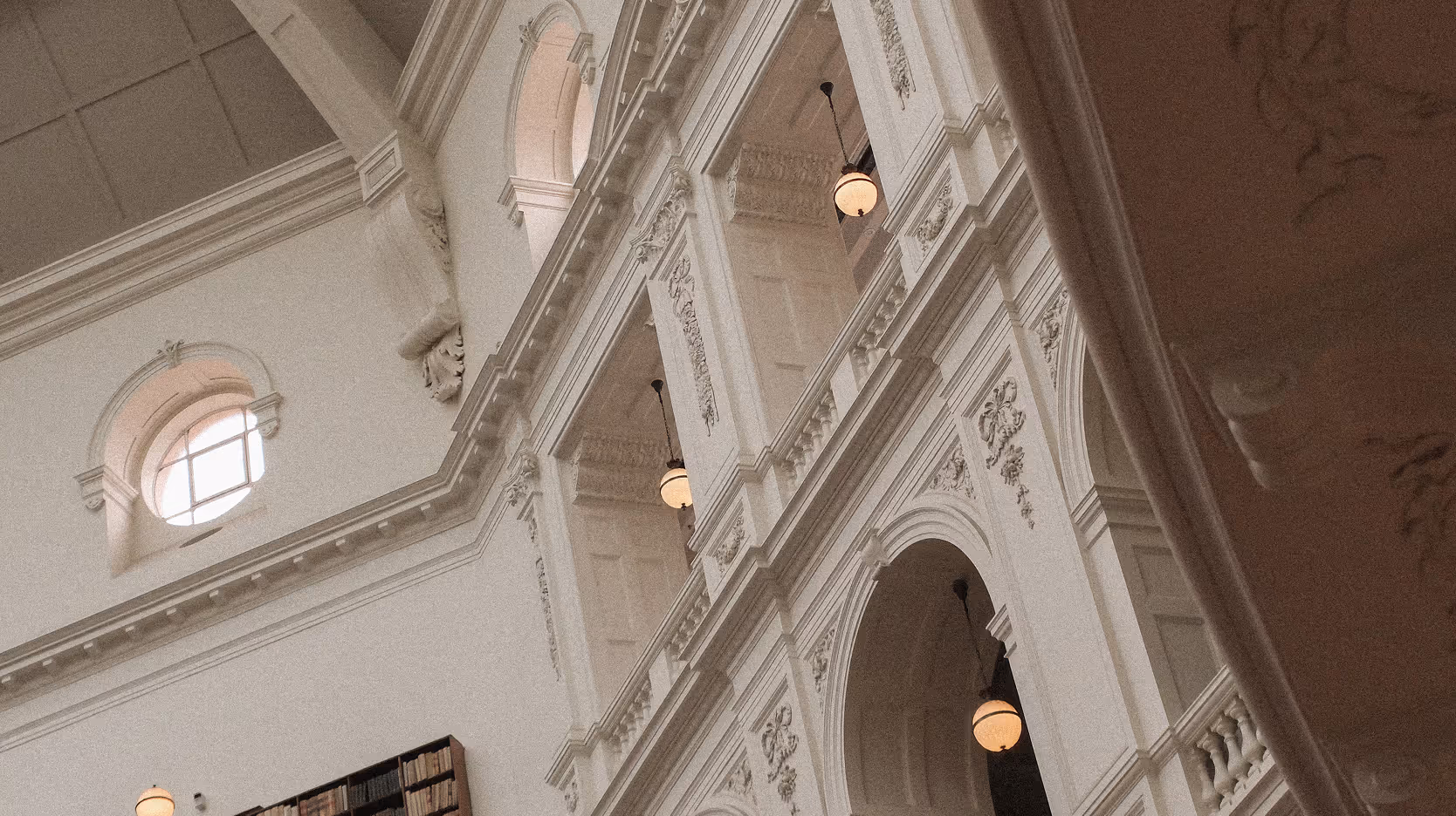 Interior view of a grand classical building with ornate white arches, decorative moldings, round windows, and hanging spherical light fixtures.
