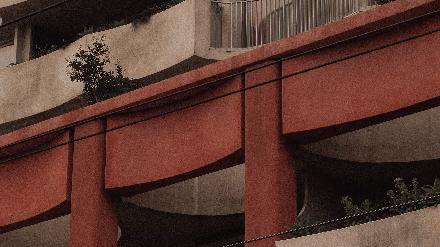 Close-up of a modern building facade featuring red architectural beams and beige concrete balconies with plants.