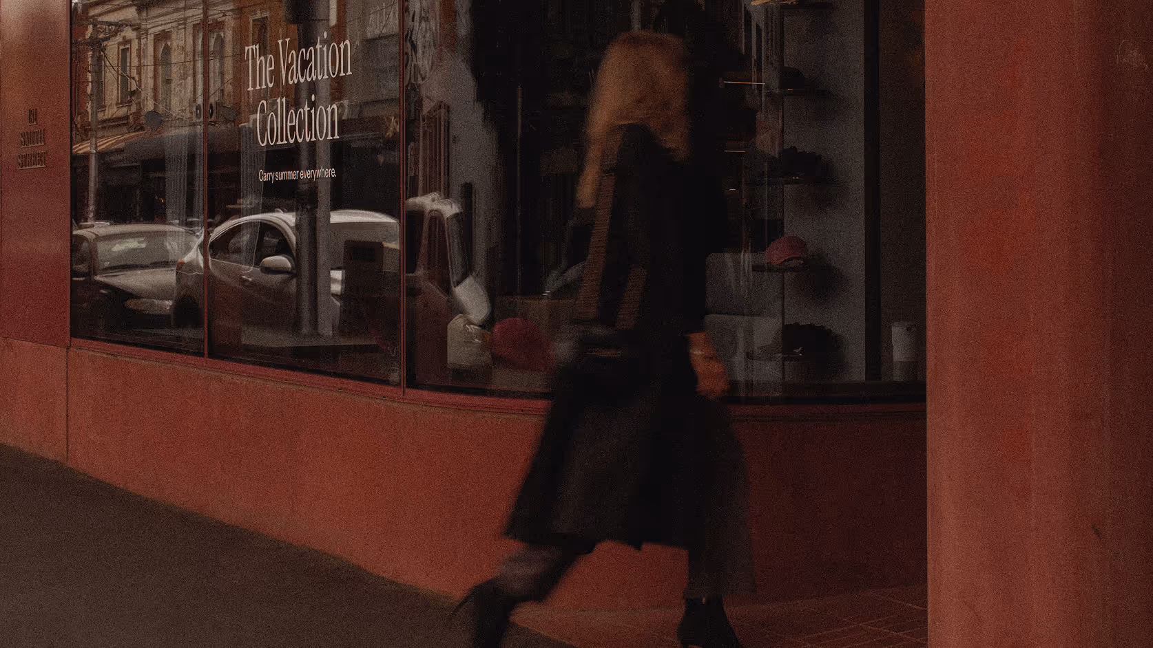 A person walking past a store window displaying a sign that reads 'The Vacation Collection' with cars reflected in the glass.