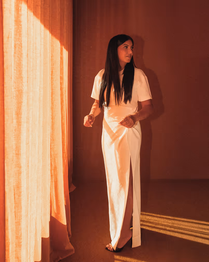 Woman in a long white dress standing barefoot near a sunlit curtain in a warm-toned room.