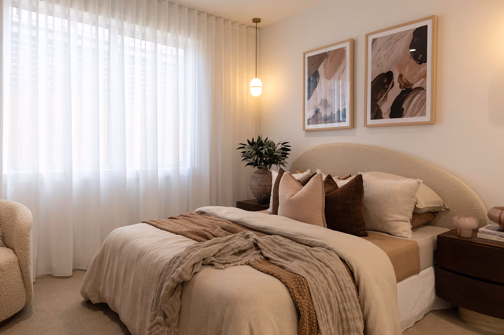 Cozy bedroom with a beige upholstered bed, layered neutral bedding, abstract wall art, and soft natural light through sheer curtains.