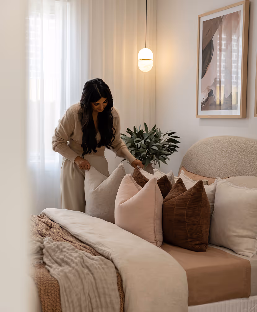 Woman arranging beige and brown pillows on a neatly made bed in a softly lit bedroom with neutral decor.