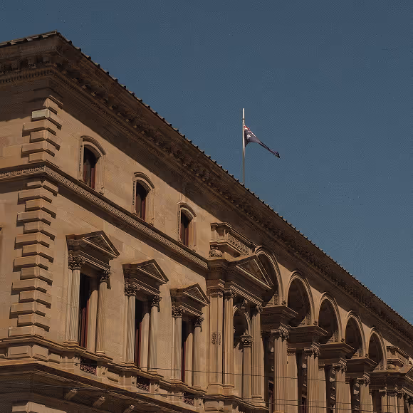 Historic sandstone building with classical architectural details and an Australian flag flying on the roof against a clear blue sky.