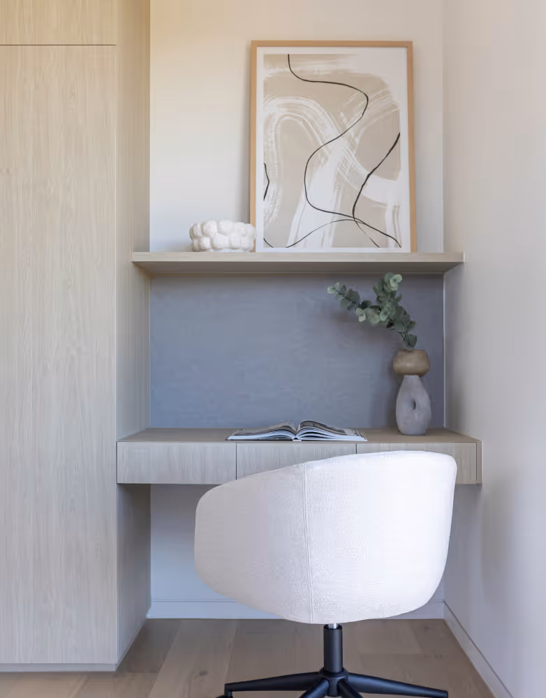 Minimalist workspace with a white swivel chair, light wood desk with open book, abstract art, and a vase with green foliage.