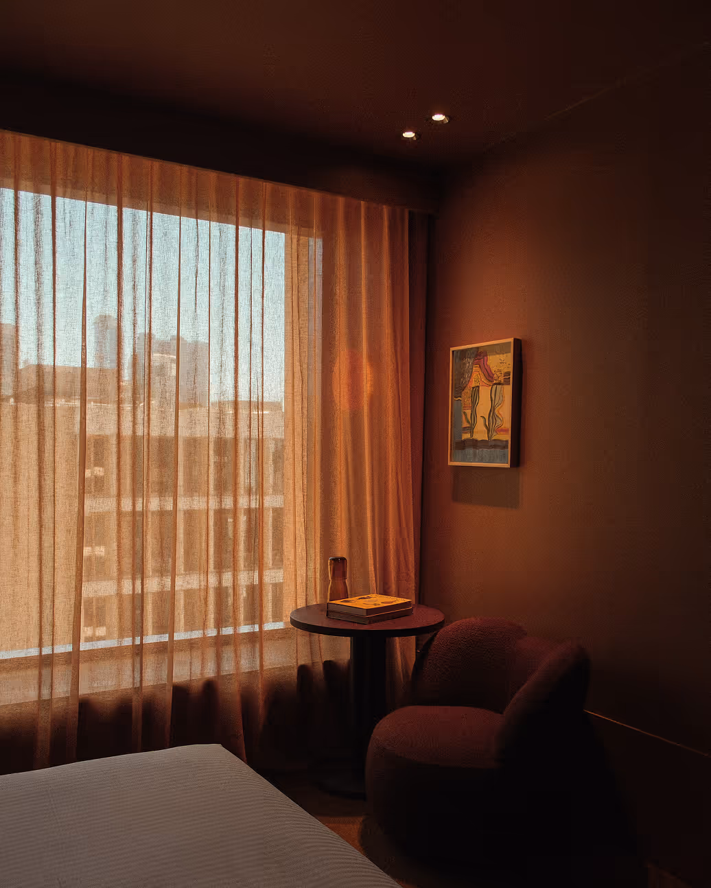 Cozy corner of a dimly lit room with sheer curtains covering large window, a small round table holding a book and a glass, next to a plush chair and a framed abstract painting on the wall.