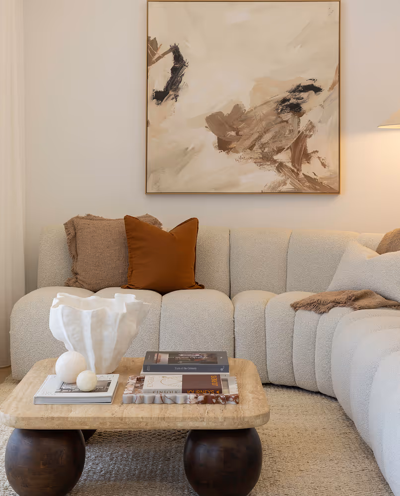 Modern beige sectional sofa with brown and beige pillows and a coffee table displaying books and decorative white ceramic pieces, beneath an abstract beige and brown painting.