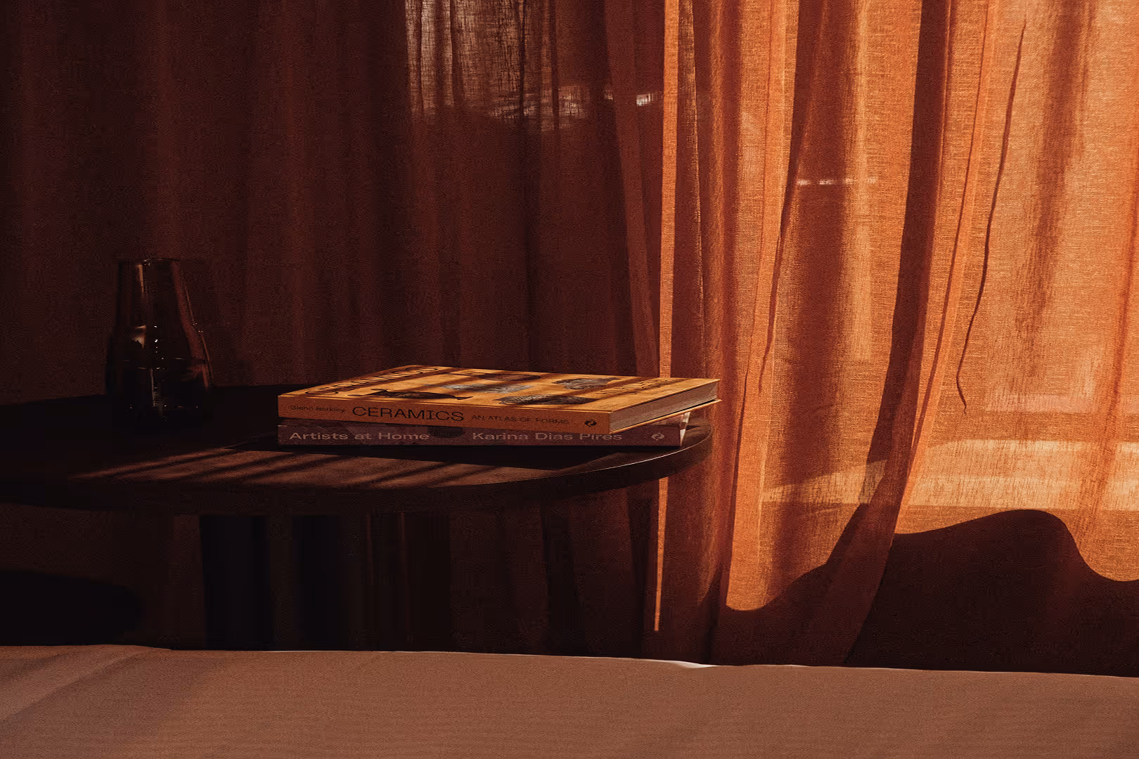 Two books stacked on a round wooden table with dark brown curtains in warm light.