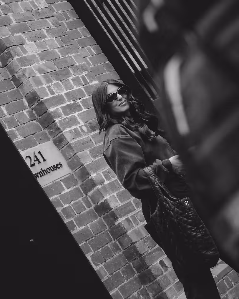 Woman wearing sunglasses and a dark jacket leaning against a brick wall next to a sign reading '241 Townhouses' in black and white.