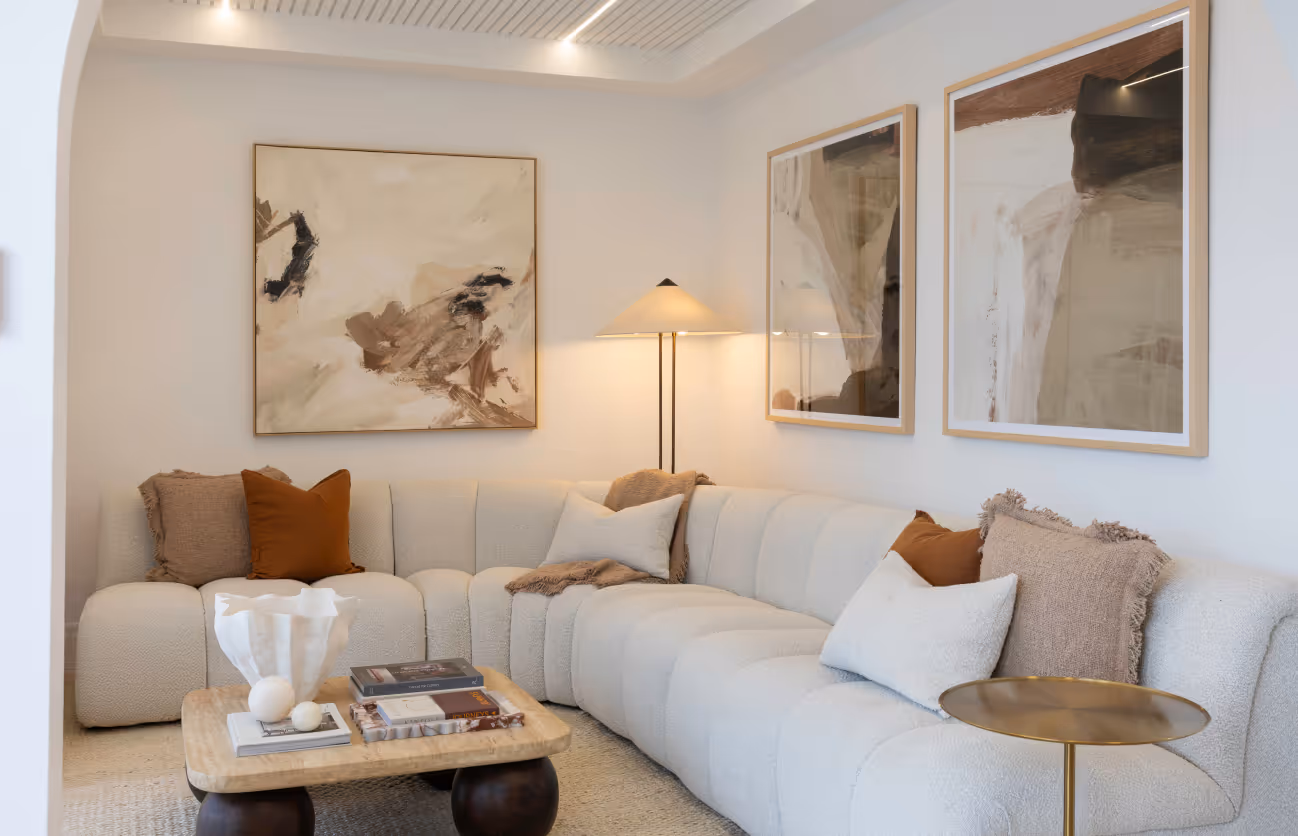 Modern living room with a white sectional sofa adorned with beige, brown, and white pillows, a wooden coffee table with books and decor, a floor lamp, and abstract art paintings on white walls.