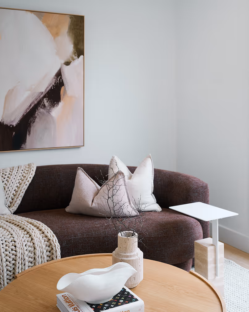 Modern living room with a curved dark brown sofa, white cushions, a chunky knit blanket, and a round wooden coffee table holding a white decorative bowl and a textured vase with branches.